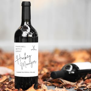 Peut inclure: Une bouteille de vin avec une étiquette blanche qui dit "Pairs well with a great Hockey Manager. Thank you for all you do." L'étiquette comporte également une illustration en noir et blanc de deux bâtons de hockey croisés.