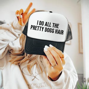 May include: Black and white trucker hat with the text "I DO ALL THE PRETTY DOGS HAIR" in bold, black letters. The hat has a white front panel and a black mesh back. The brim is black.