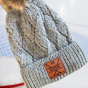 May include: Grey cable knit beanie with a brown leather patch that says "15 EIDEN" with a hockey stick and puck graphic.