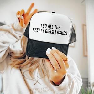 Puede incluir: Gorra de camionero en blanco y negro con el texto "I DO ALL THE PRETTY GIRLS LASHES" en negro. La gorra tiene un panel frontal blanco y una visera y parte trasera de malla negras.