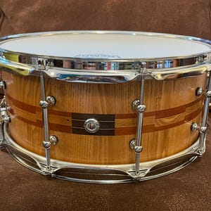 May include: A wooden snare drum with chrome hardware. The drum has a natural wood finish with red and black stripes. The drum is 14 inches in diameter.
