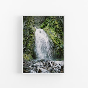May include: A close-up view of a waterfall cascading down a mossy rock face. The water is white and foamy, and the rocks are dark and wet. The surrounding foliage is lush and green.