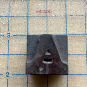 May include: A dark gray letterpress printing block featuring the capital letter "A". The block is square and has a slightly worn appearance, indicating use. The letter "A" is clearly defined with a small triangular serif at the top.