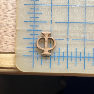 May include: A small, wooden Phi symbol, approximately 0.75 inches tall, is centered on a clear, grid-marked surface. The symbol is a light brown color, with a classic design. The grid has blue lines and is used for measurement.