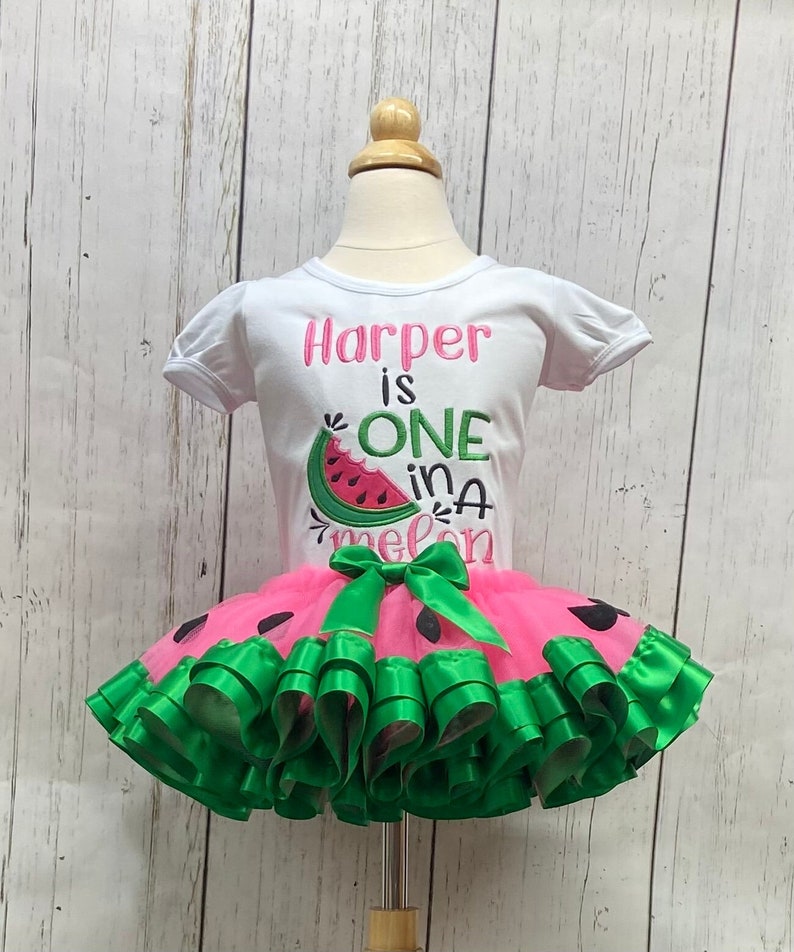 Op de afbeelding: Een wit T-shirt met de tekst "Harper is ONE in a melon" en een watermeloenafbeelding. Het shirt wordt gecombineerd met een roze tule tutu met groene lintversiering en zwarte watermeloenzaadaccenten.