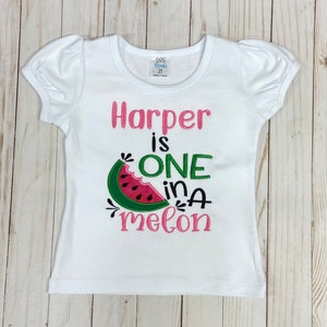 Op de afbeelding: Wit T-shirt met korte mouwen met roze en groene borduurwerk met de tekst "Harper is one in a melon" met een watermeloenafbeelding.