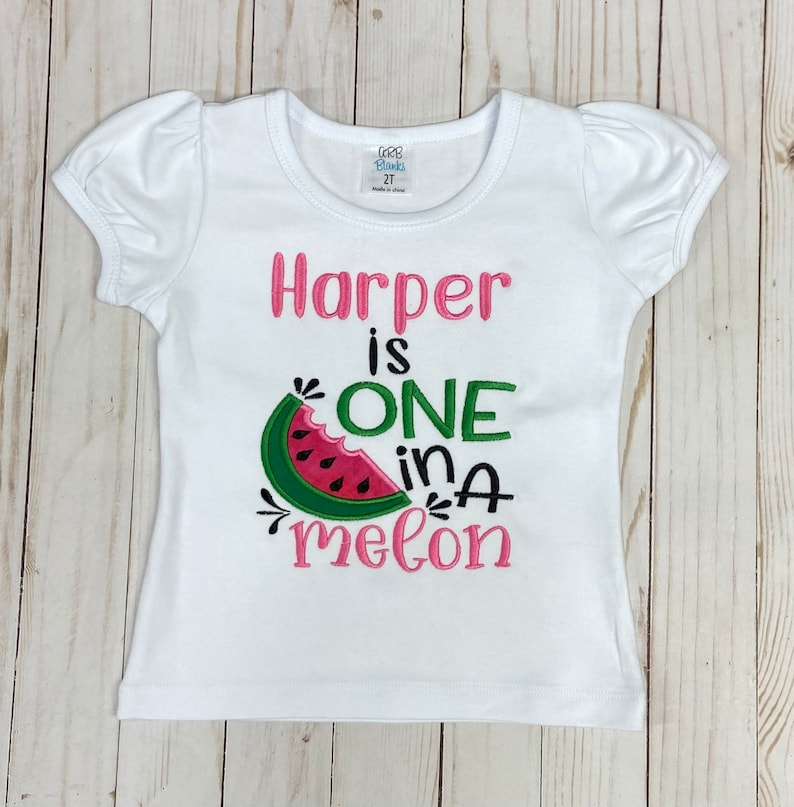 Op de afbeelding: Wit T-shirt met korte mouwen met roze en groene borduurwerk met de tekst "Harper is one in a melon" met een watermeloenafbeelding.