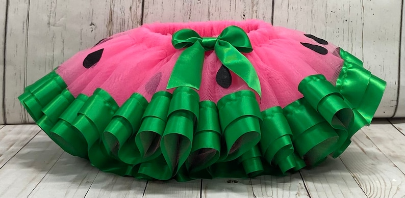 Op de afbeelding: Een roze tule tutu met een groene lintversiering en een groene strik. De tutu is versierd met zwarte watermeloenpitten.