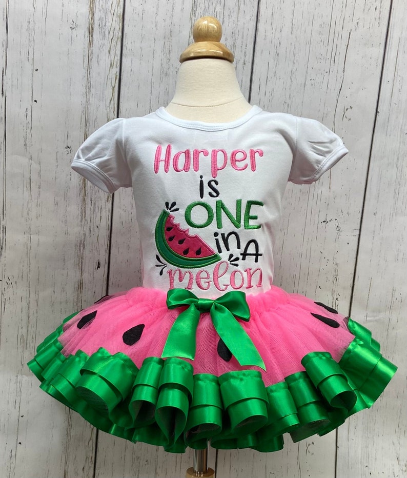 Op de afbeelding: Een wit T-shirt met de tekst "Harper is ONE in a melon" en een watermeloenafbeelding. Het shirt wordt gecombineerd met een roze tule tutu met groene lintversiering en een groene strik.