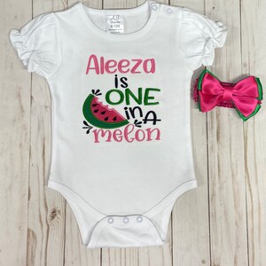 Op de afbeelding: Witte romper met roze en groene borduurwerk met de tekst "Aleeza is one in a melon". Een roze strik hoofdband is inbegrepen.