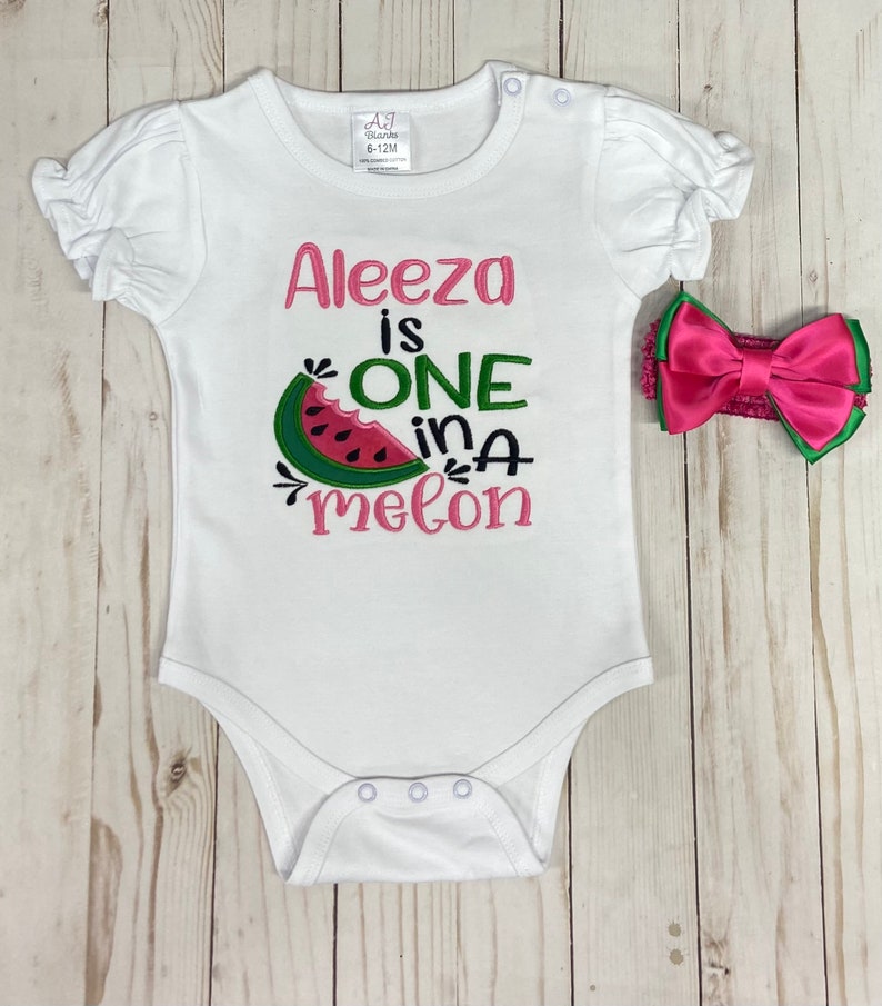 Op de afbeelding: Witte romper met roze en groene borduurwerk met de tekst "Aleeza is one in a melon". Een roze strik hoofdband is inbegrepen.