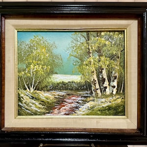 May include: Oil painting of a landscape scene with a stream, trees, and a bright sky. The painting is framed in a black and gold frame. The artwork depicts a serene outdoor scene with a stream flowing through a forest.