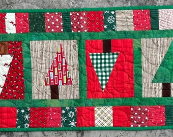 May include: A green, red and white patchwork table runner with a Christmas tree theme. The runner features a variety of fabrics with different patterns, including plaid, polka dots and snowflakes. The runner is approximately 46 cm wide and 122 cm long.