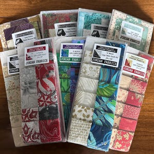 May include: A collection of precut fabric squares for quilting, featuring various patterns and colors. Each package is labeled with "Precision Cut Jordan Fabrics" and includes kits for log cabin quilts and table runners. The dimensions of the quilt kits are 29 x 39 inches.