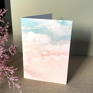 Könnte beinhalten: Eine gefaltete Grußkarte mit einem Aquarelldesign eines pastellfarbenen Himmels. Der Himmel geht von Hellblau oben nach Rosa unten über, mit flauschigen weißen Wolken und einem kleinen weißen Herzen. Die Karte ist geöffnet und zeigt ein leeres Innere.