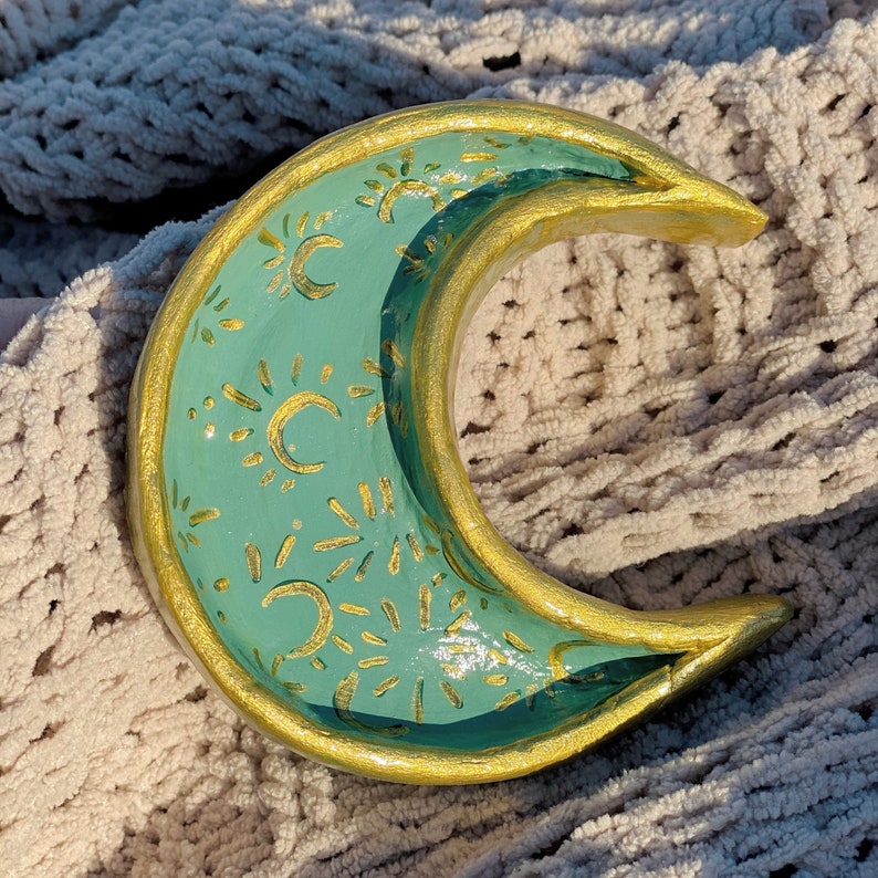 May include: A crescent moon shaped ceramic dish with a teal glaze and gold accents. The dish is decorated with a repeating pattern of suns and moons.