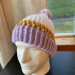 May include: A purple and white knitted beanie with a white pom pom on top. The beanie has a band of gold and white yarn with pink and blue beads.