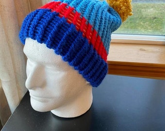 May include: Blue, red, and light blue knitted beanie with a yellow pom pom on top.