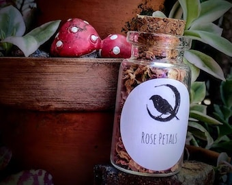 May include: A small glass jar filled with dried rose petals. The jar has a cork lid and a white label that says "Rose Petals". A black silhouette of a bird is on the label.