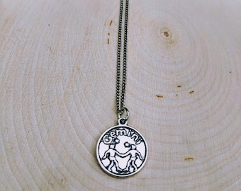 May include: A silver necklace with a round pendant featuring the Gemini zodiac symbol. The pendant has two figures holding hands and the word "Gemini" written around the edge.