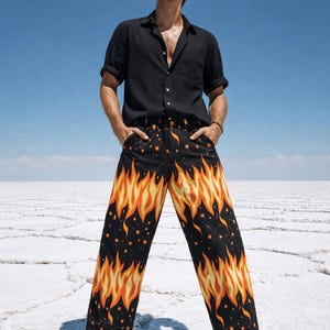 May include: Black button-down shirt and black sandals. The pants have a black background with a flame design in orange and yellow, and small star patterns. The person is standing on a white surface under a blue sky.