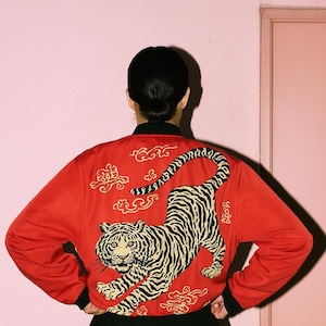 Puede incluir: Chaqueta bomber roja con cuello y puños negros. La parte trasera de la chaqueta presenta un gran diseño de tigre en negro y beige, con detalles dorados. La chaqueta se combina con una falda plisada negra.