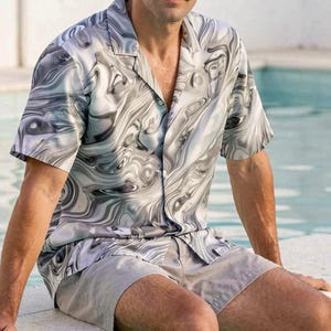 Pode incluir: Um conjunto de camisa de manga curta e calções a condizer. A camisa apresenta um padrão cinzento e branco rodopiante. Os calções são de cor cinzento claro lisa. A pessoa está sentada na borda de uma piscina.