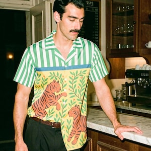 May include: A short-sleeve button-down shirt featuring a green and white vertical striped pattern on the upper section and a yellow lower section with a repeating tiger and foliage print, paired with solid black trousers and a brown leather belt.