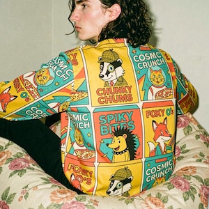 May include: A long-sleeve shirt featuring a vibrant, repeating grid pattern of colorful cereal box illustrations. The graphic print includes cartoon characters like a fox, a hedgehog, and a badger, with text reading Chunky Chums, Spiky Bites, and Foxy O's.