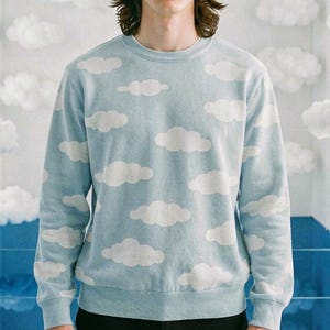May include: A light blue sweatshirt with a white cloud pattern. The crew neck sweatshirt is worn by a person standing in front of a wall decorated with fluffy white clouds. The person is wearing black pants.