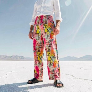 May include: A person in a white shirt and patterned trousers. The trousers have a colourful, abstract design with yellow figures and pink hues. The outfit is completed with black sandals. The background is a bright, sunny landscape with mountains in the distance.