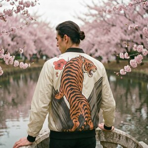 Könnte beinhalten: Cremefarbene Bomberjacke mit einer detaillierten Illustration eines Tigers mit orangefarbenen und schwarzen Streifen. Die Jacke hat auch florale Akzente und einen schwarzen Kragen. Die Jacke wird im Freien in der Nähe eines Gewässers getragen.