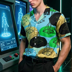 Op de afbeelding: Een overhemd met korte mouwen met een geometrisch patroon in zwart, goud, groen en blauw. Gedragen met een zwarte broek en een zwarte riem. De achtergrond toont futuristisch ogende schermen en een figuur in een deuropening.