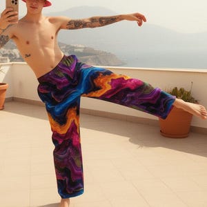 May include: A person wearing colorful patterned pants, a pink bucket hat, and holding a phone. The pants feature a vibrant design with swirls of blue, purple, and orange. The hat has a small embroidered design. The background shows a coastal view.