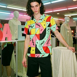 May include: A short-sleeve button-down shirt featuring a vibrant geometric pattern in yellow, pink, teal, and black with squiggly lines and dots. The shirt has a notched collar and is paired with solid black shorts, creating a bold, retro-inspired look.