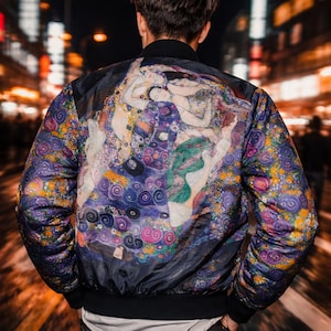 Puede incluir: Chaqueta bomber negra con un diseño artístico y colorido en la espalda. La obra de arte presenta figuras en tonos morados, azules, verdes y dorados. La chaqueta tiene cuello y puños negros. La persona lleva vaqueros negros.