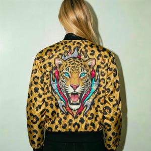 May include: A bomber jacket featuring a tan and black leopard print pattern. The back displays a large, detailed illustration of a snarling leopard head with bright blue eyes, surrounded by stylized red and silver flames with sparkling accents.
