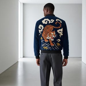May include: Navy blue bomber jacket with a detailed embroidered tiger design on the back. The tiger is orange and black, set against a backdrop of white clouds and yellow accents. The jacket has black cuffs and a black waistband.
