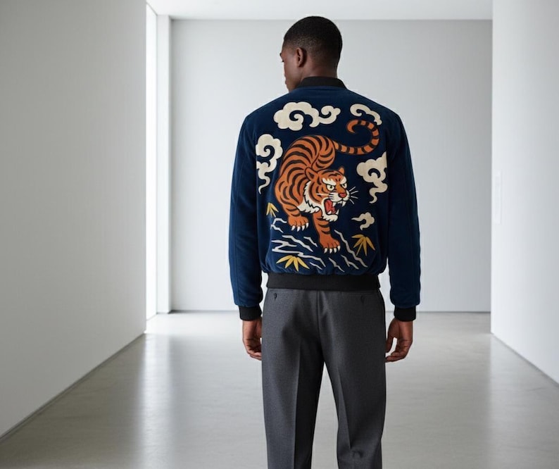 May include: Navy blue bomber jacket with a detailed embroidered tiger design on the back. The tiger is orange and black, set against a backdrop of white clouds and yellow accents. The jacket has black cuffs and a black waistband.