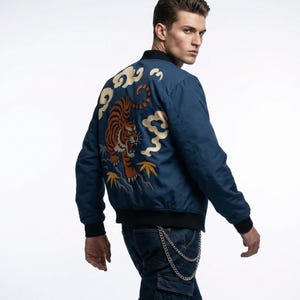 May include: A navy blue bomber jacket with a detailed embroidered tiger design on the back. The tiger is orange and black, set against a backdrop of gold clouds and yellow leaves. The jacket has black trim and cuffs.