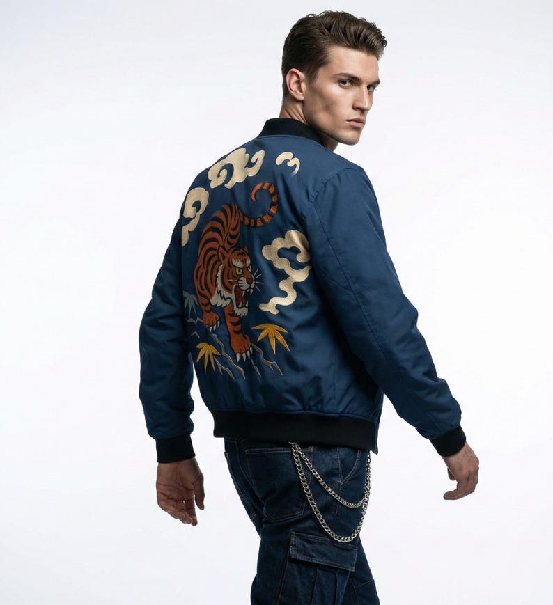 May include: A navy blue bomber jacket with a detailed embroidered tiger design on the back. The tiger is orange and black, set against a backdrop of gold clouds and yellow leaves. The jacket has black trim and cuffs.