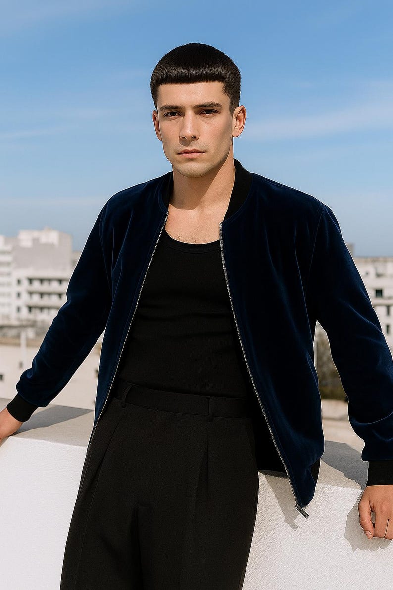 May include: A navy blue bomber jacket with a black shirt and black pants. The jacket has a zipper closure and ribbed cuffs. The outfit is worn by a person standing outside with a blue sky background.