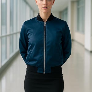 May include: A navy blue bomber jacket with a black collar and cuffs, featuring a silver zipper. The jacket is paired with a black skirt. The woman is standing in a hallway with large windows.