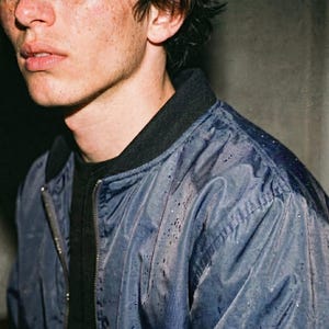 May include: A close-up of a person wearing a blue bomber jacket with a black collar and a black shirt underneath. The jacket has a zipper closure and appears to be slightly wet. The person has freckles and dark hair.