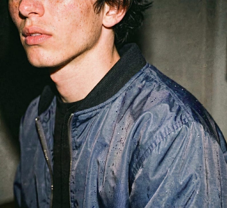 May include: A close-up of a person wearing a blue bomber jacket with a black collar and a black shirt underneath. The jacket has a zipper closure and appears to be slightly wet. The person has freckles and dark hair.