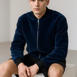May include: A navy blue velvet bomber jacket with a silver zipper and black ribbed cuffs. The jacket is paired with black shorts. The model is sitting on a white cube.