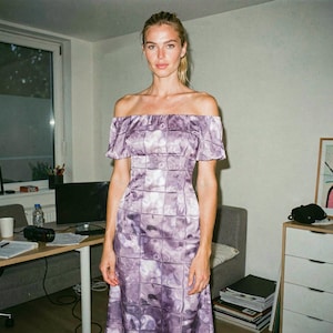 Puede incluir: Vestido lila con hombros descubiertos y estampado de cuadros. El vestido tiene mangas cortas y largo midi. La tela tiene un acabado brillante. La modelo está de pie en una habitación con un escritorio y un ordenador.