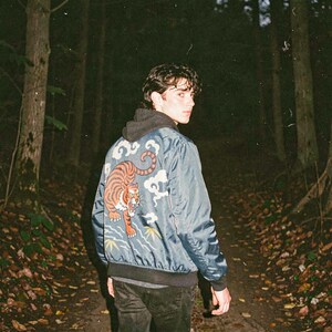 May include: A blue bomber jacket with a tiger design on the back. The jacket features a black collar and cuffs, and the tiger is orange and black. The background is a forest with a path.