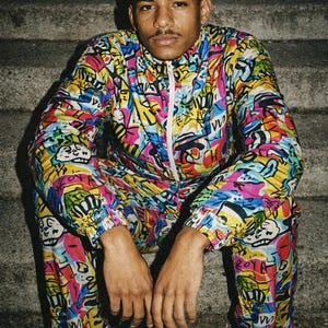 May include: A person wearing a colorful tracksuit with a white zipper and white sneakers. The tracksuit features a vibrant pattern of various shapes and colors, including pink, yellow, blue, and green. The person is sitting on concrete steps.