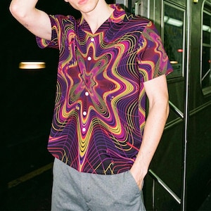 Puede incluir: Camisa de manga corta con un vibrante estampado en espiral en tonos morados, amarillos y rosas. La camisa tiene un cuello clásico y botones blancos. Se combina con pantalones grises.